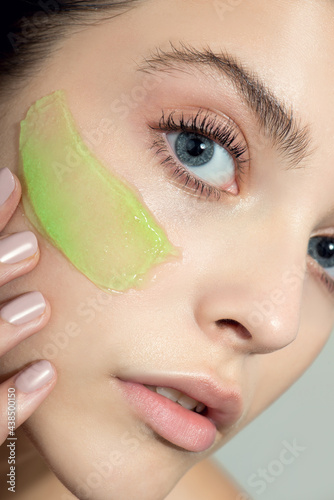 Young woman applying green gel face cream or facial mask at her face. Beauty model with perfect fresh skin and long eyelashes cares about her skin at home. Close up, selected focus
