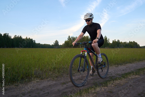 A male cyclist rides a mountain bike on a dirt road. Dressed in a bicycle uniform and a helmet. Evening endurance training. Sports and healthy lifestyle.