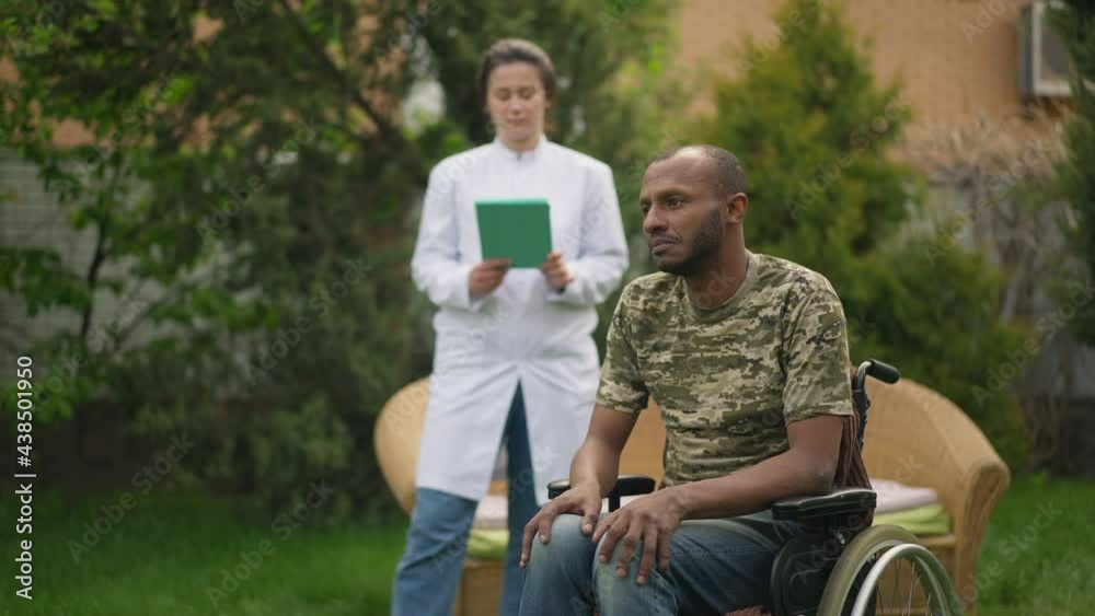 Sad desperate disabled man in wheelchair looking away as doctor nurse standing at background outdoors. Portrait of hopeless African American military patient and Caucasian caregiver in nursing home Sad desperate disabled man in wheelchair looking away as doctor nurse standing at background outdoors. Portrait of hopeless African American military patient and Caucasian caregiver in nursing home