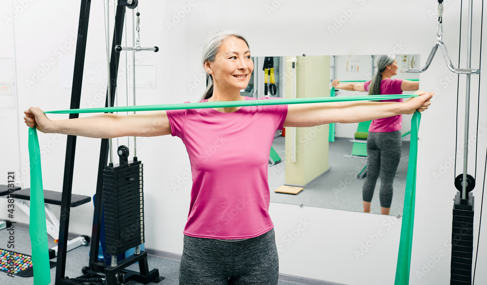Fototapeta premium Patient woman doing exercises for recovery arms after injury using stretching resistance band at a rehab center