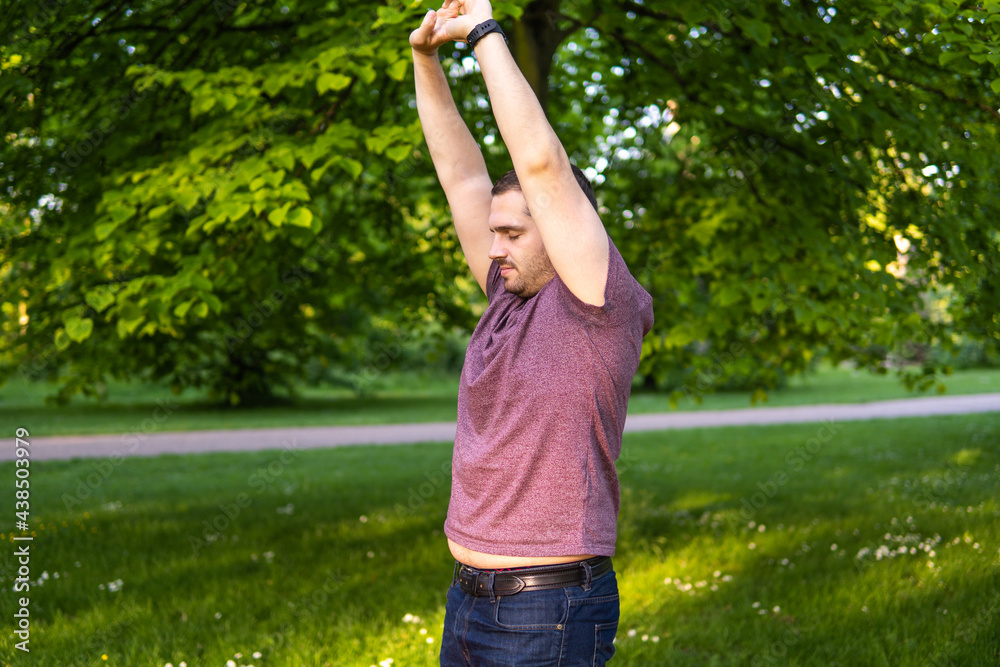 Photo of a young and attractive man in the park streching with his arms up to the sky and his eyes closed. Alone in nature during a summer day. Yoga pose