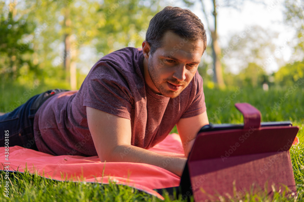 Photo of a young and attractive man sitting in the park on a towel enjoying a sunny day outside in the nature and checking his tablet. Electronic device, technology