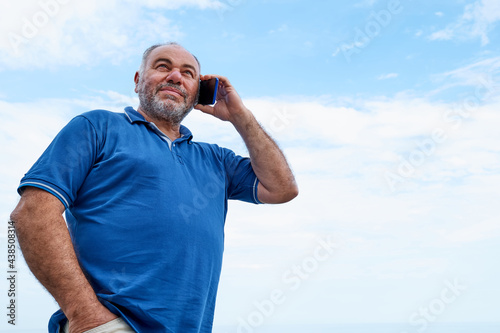 Handsome middle-aged bearded man talking on cell phone while walking on beach with sea in background. Communication and connectivity concept.