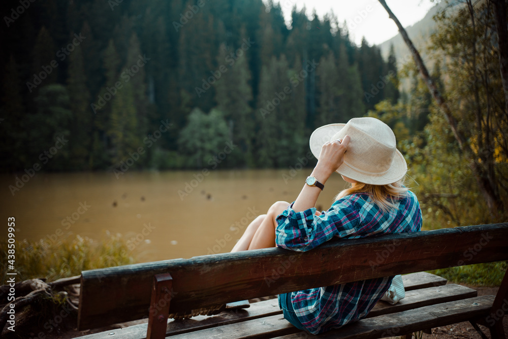 Young woman in a denim shirt and hat, sitting on a bench, admires the lake landscape, the pine forest and the relief of the mountains in the background