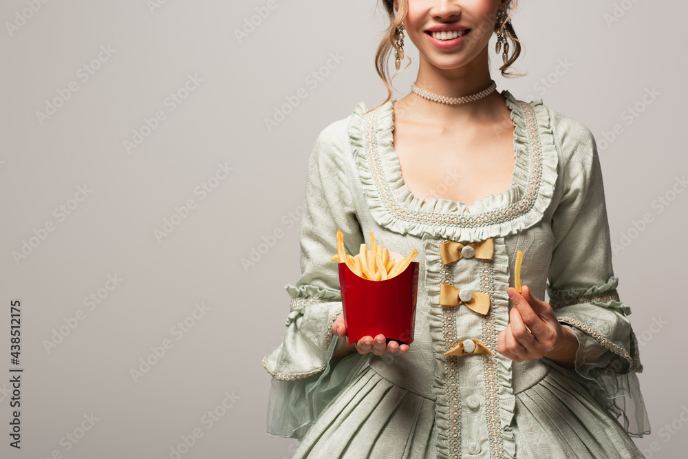 cropped view of smiling woman in retro dress holding french fries isolated on grey