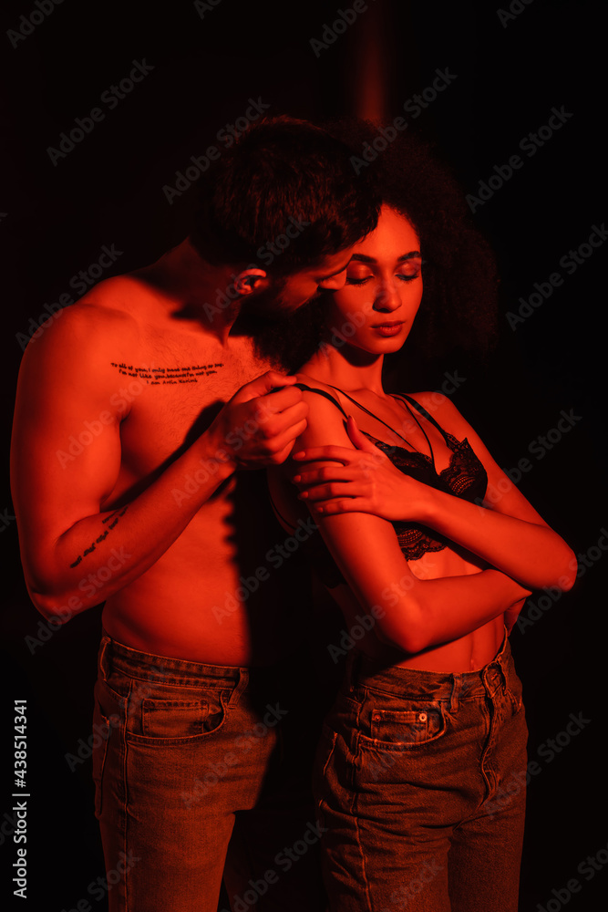 Muscular man taking off bra from sensual african american woman in red light on black background
