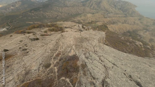 Mountain bare top with brown rocky cliffs and dense thickets on hilly landscape against blue sea under sunlight aerial cinematic view. Flight from the top at high speed dive down on sport fpv drone photo