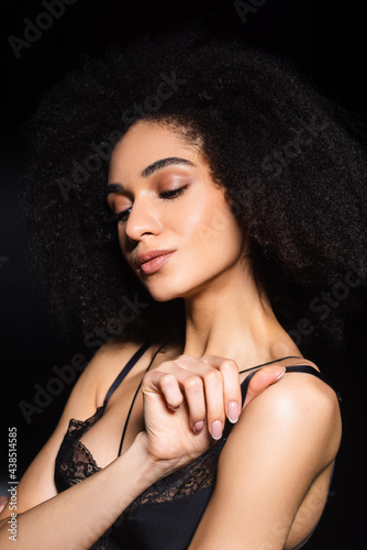 Seductive african american woman holding strap of lace bra isolated on black
