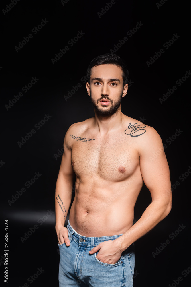 Fototapeta premium Tattooed man in jeans holding hands in pockets on black background
