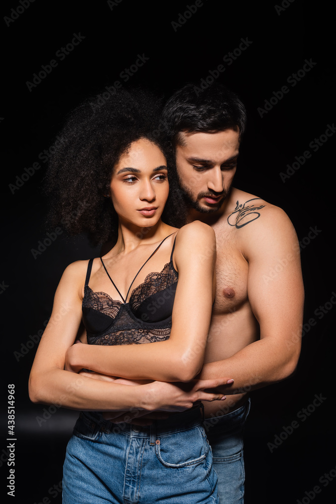 Muscular man embracing african american girlfriend in bra and jeans on black background