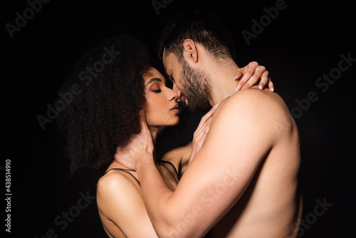 Side view of shirtless man kissing african american woman isolated on black