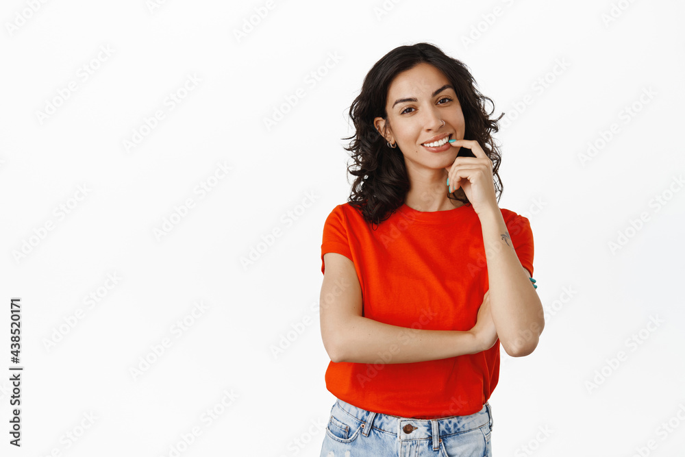 Stylish brunette girl with curly hairstyle, red t-shirt, biting finger coquettish while thinking, making choice or decision, standing over white background Stylish brunette girl with curly hairstyle, red t-shirt, biting finger coquettish while thinking, making choice or decision, standing over white background