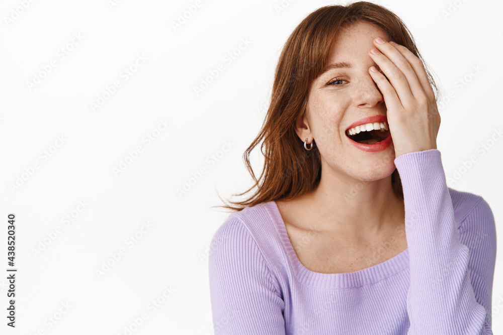 Fototapeta premium Candid young woman covering half of face, laughing and smiling white teeth, showing glowing facial skin, standing in blouse against white background