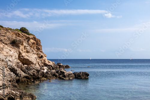Wild mediterranean sea with rocky cliffs shore, sailing boat in distance and blue clear water. Travel Greece near Athens. Summer nature scenic view