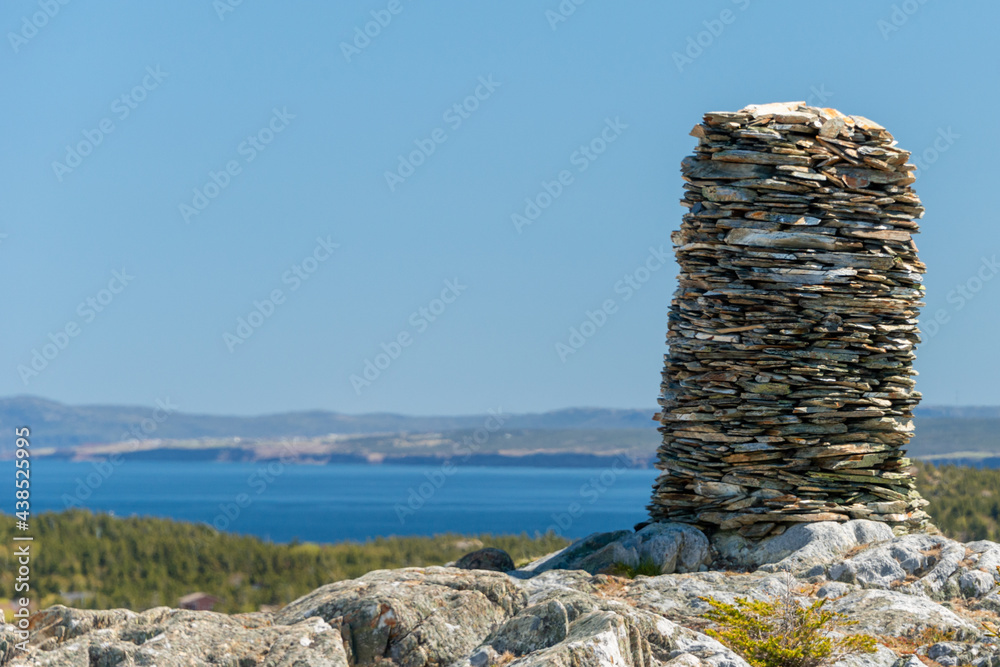 A traditional trail marker called a cairn. The large mass of rocks is stacked in a circle forming a cylinder shape inukshuk. The rocks are piled on the top of a mountain with blue sky and ocean. 