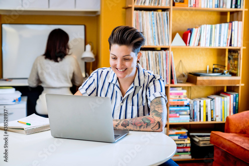 Smiling woman working on laptop while wife works behind her