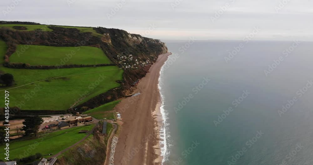 4K Smooth Aerial shot descending towards the oceans edge and glimpsing the mountains, cottages and rolling hills of Branscombe Beach