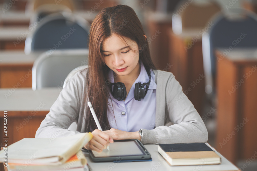 Stock photo of a young teenage woman Asian college student in student uniform studying and writing on digital tablet in a university classroom