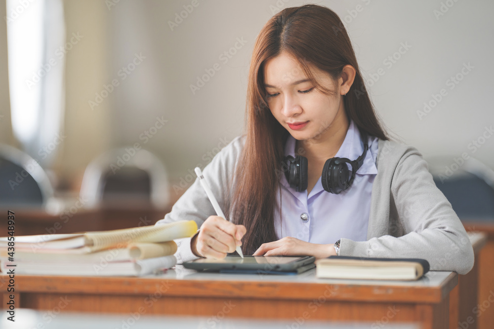 Stock photo of a young teenage woman Asian college student in student uniform studying and writing on digital tablet in a university classroom Stock photo of a young teenage woman Asian college student in student uniform studying and writing on digital tablet in a university classroom