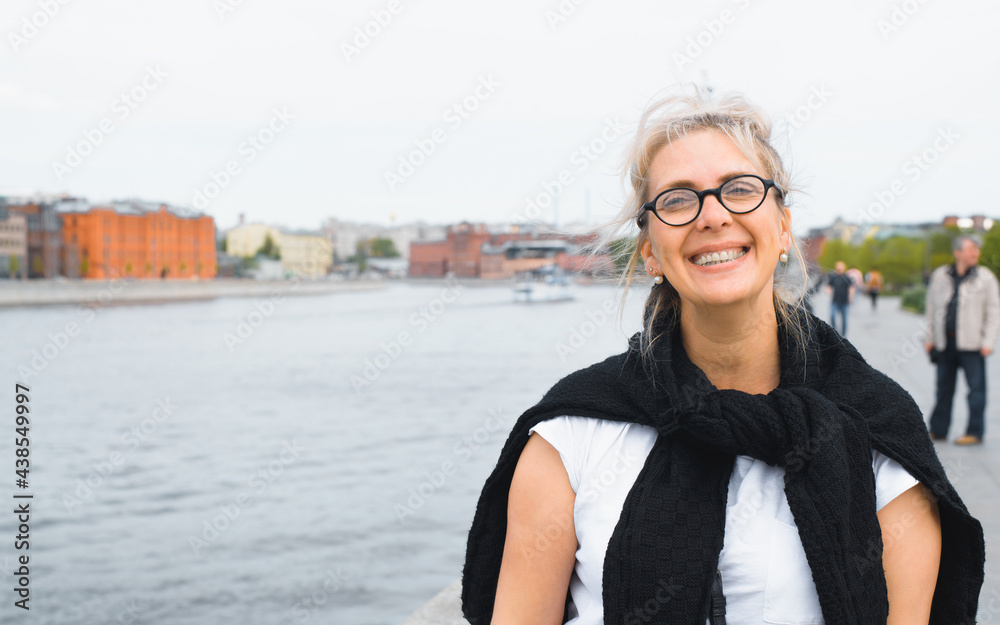 Portrait of happy woman standing on the river embankment and looking at the camera. Smiling tourist woman, blurred background river and cityscape. Concept for tourist trip, vacation, lifestyle.