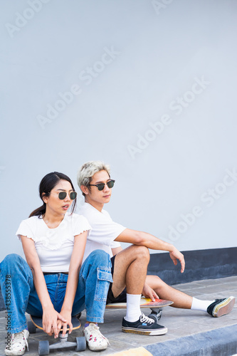 Asian handsome man and cute woman sitting on skateboards, wear sunglasses, look at right side in front of cool wall with copy space on wall in daylight time, summer holiday