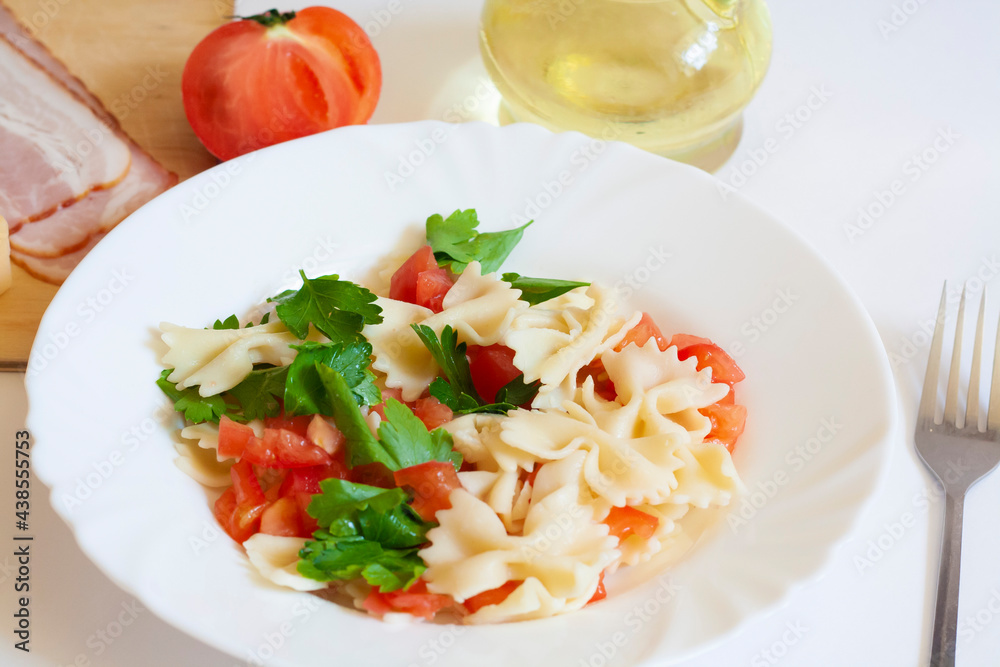 Pasta farfalle with tomatoes and herbs in a plate, fork, bacon, cheese, tomato and oil in a jug on a white background Pasta farfalle with tomatoes and herbs in a plate, fork, bacon, cheese, tomato and oil in a jug on a white background