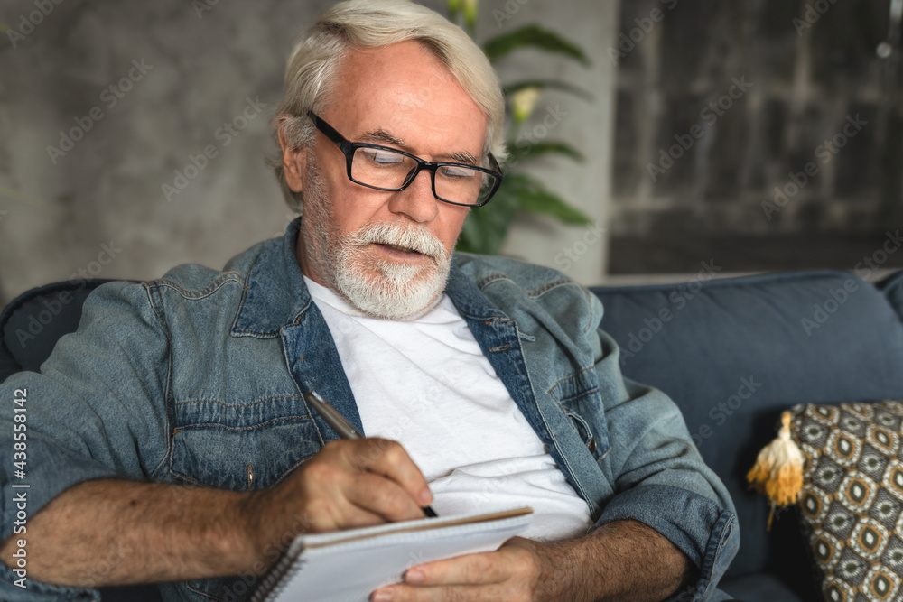 Senior man taking notes in notebook while sitting on the couch, clever gray-haired elderly man writing down thoughts and ideas in notebook Senior man taking notes in notebook while sitting on the couch, clever gray-haired elderly man writing down thoughts and ideas in notebook