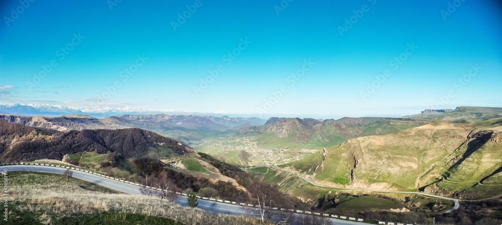 Aerial panoramic landscape view over Gum-bashi mountain pass, with green grass on cliffs, winding road on hills, Сaucasus mountian range and Elbrus mountain. Karachay-Cherkessia
