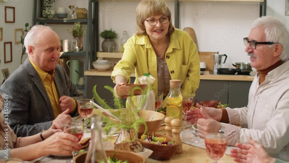 Cheerful senior woman passing dish to friends and sitting at table while hosting holiday dinner party at home Cheerful senior woman passing dish to friends and sitting at table while hosting holiday dinner party at home