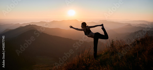 Sporty young woman performing yoga pose on grassy hill with orange morning sky on background. Female person standing on one leg and doing yoga exercise outdoors. Panoramic view.