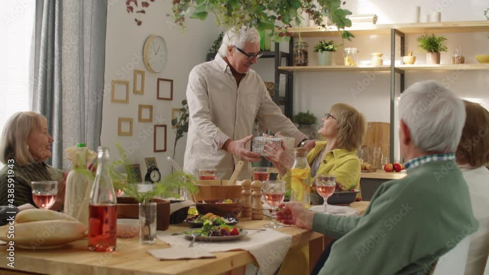 Joyous elderly man giving gift box to happy wife, saying wishes, kissing and hugging her while smiling friends clapping hands at birthday dinner Joyous elderly man giving gift box to happy wife, saying wishes, kissing and hugging her while smiling friends clapping hands at birthday dinner