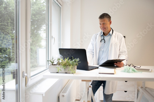 handsome doctor with white coat and blue shirt and stethoscope stands behind high table and works on laptop and has web conference or meeting
