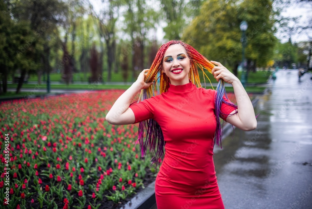Cute girl with multi-colored African braids and bright expressive makeup in a tight red dress is spinning in the park enjoying the arrival of spring