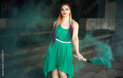 Cute girl in a green dress with African braids and colorful makeup posing with bright green smoke on the streets of a warm spring city
