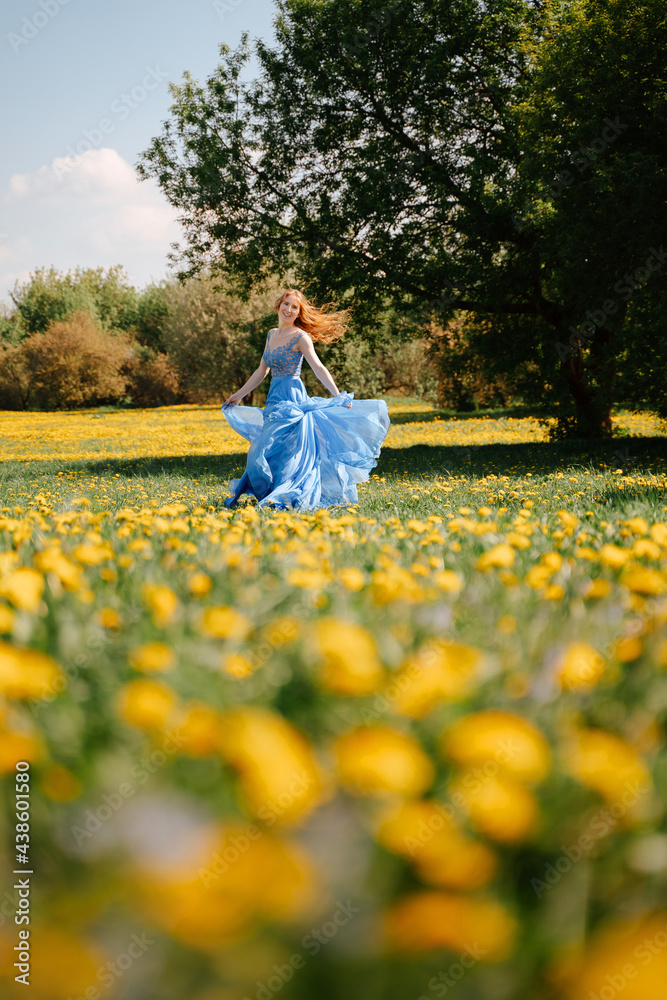 Fototapeta premium A happy fairy-tale girl in an airy blue dress runs joyfully along the green lawn in the park. Blooming field in yellow dandelions. Allergy free concept.