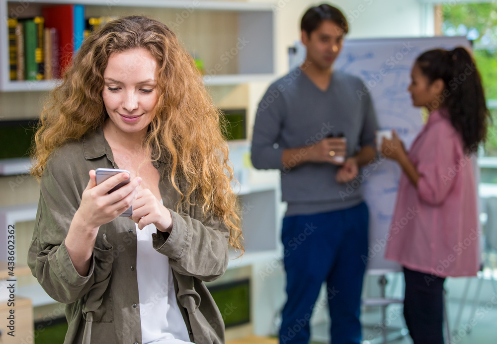 Foreground focus Business Creative Caucasian female stylish or designer holding, sending message, using mobile phone and smart standing in indoor modern office with background multiethnic colleagues. Foreground focus Business Creative Caucasian female stylish or designer holding, sending message, using mobile phone and smart standing in indoor modern office with background multiethnic colleagues.