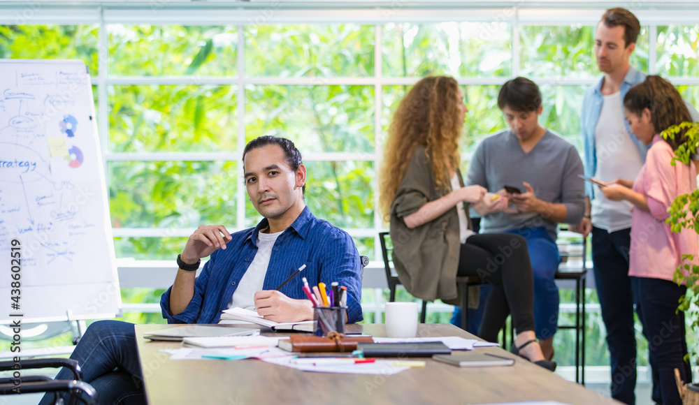 Foreground focus handsome hipster Caucasian male creative director, boss or leader sitting in comfortable modern office, smiling with confidence and profession with background of mixed race employee.
