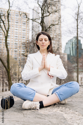 Young adult woman meditating sitting crossed leg lotus pose pray position in white jacket, blue jeans in New York city Central park. Peaceful time in busy life 
