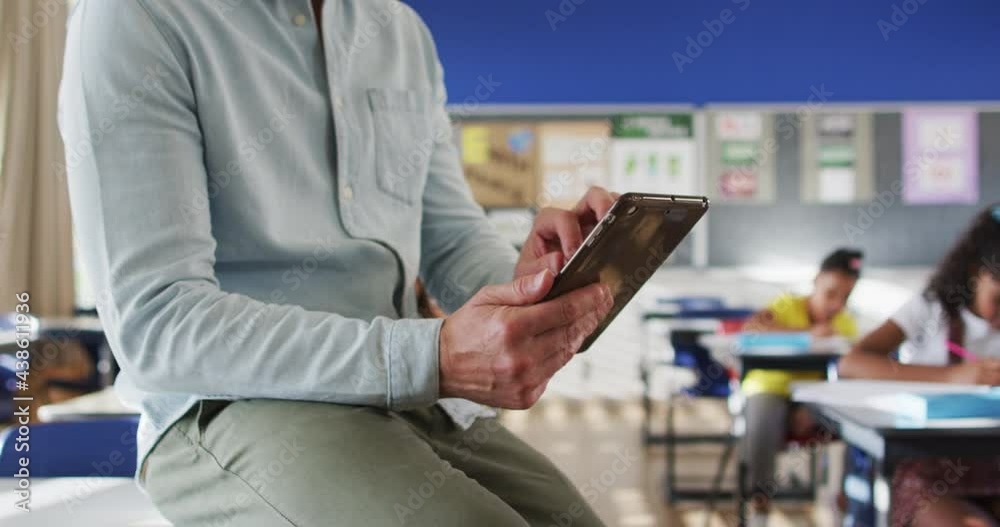 Portrait of happy caucasian male teacher in classroom with children using tablet looking at camera Portrait of happy caucasian male teacher in classroom with children using tablet looking at camera