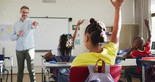 Happy caucasian male teacher in classroom with children raising hands during lesson photo