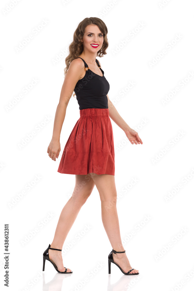 Young Woman Is Walking In Brown Suede Mini Skirt And High Heels And Looking At Camera. Side View. Young Woman Is Walking In Brown Suede Mini Skirt And High Heels And Looking At Camera. Side View.