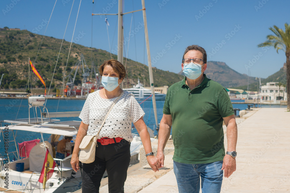 senior couple with face mask on holidays during covid19 - happy and sweet mature husband and wife enjoying retirement walking on sea harbor on sailing club