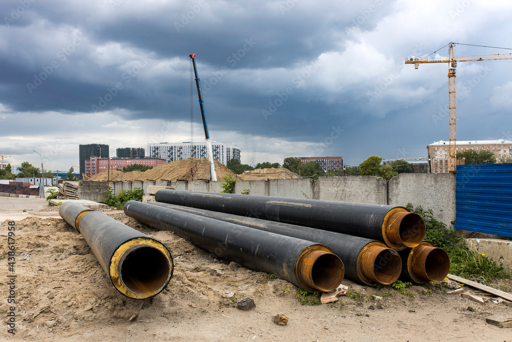 Long factory pipes lie on the ground against the backdrop of a residential area under construction Long factory pipes lie on the ground against the backdrop of a residential area under construction