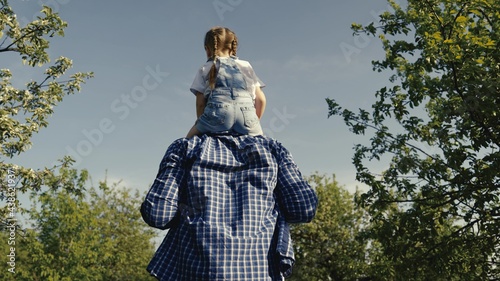 Dad plays with his daughter  carries his beloved child on his shoulders in summer park. A happy family. Father walks with baby  kid loves daddy  family walk in nature  outdoors. Parent  childhood