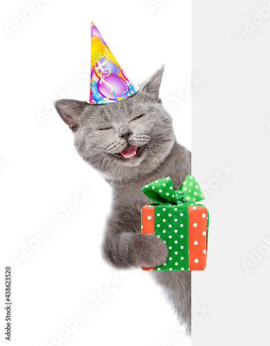Fototapeta Naklejka Na Ścianę i Meble -  Happy cat  wearing party cap holds gift box and looks from behind empty white banner. isolated on white background