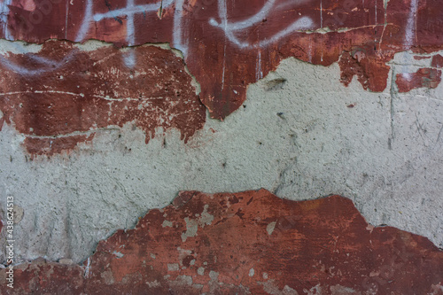 old layer of red cement plaster and fragments of vandalism of white letters falls off the wall.