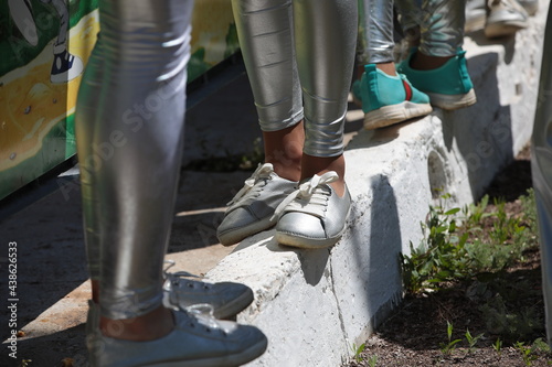 Close-up of the legs of teenagers in sports shoes and silver leggings standing by the fence on the curb in a narrow confined space.The image of the limbs of a group of people outdoors.Lower body