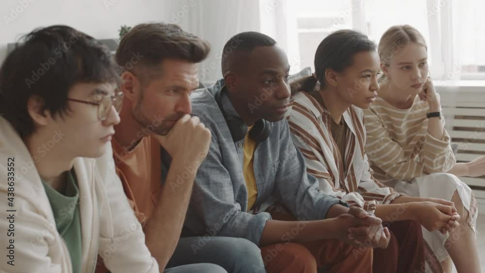 Medium long of five young multiethnic people sitting on couch in living room at home, watching invisible TV, Mixed-Race woman talking to Caucasian friend Medium long of five young multiethnic people sitting on couch in living room at home, watching invisible TV, Mixed-Race woman talking to Caucasian friend