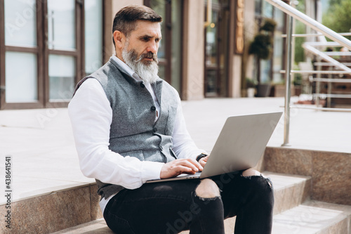 Senior man stylish man with gray beard spends time outdoors in laptop.