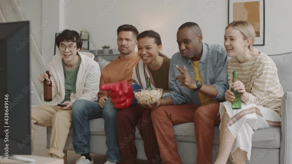 Steadicam of multiethnic young people sitting on couch in living room, drinking beer and eating snacks, watching favourite sports team winning on television Steadicam of multiethnic young people sitting on couch in living room, drinking beer and eating snacks, watching favourite sports team winning on television